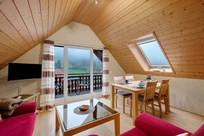 A cozy loft bedroom with a large window, a flat-screen TV, and a dining area with wooden furniture.