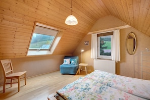 A bedroom with a sloped ceiling, a bed, a chair, a small table, a window, and a radiator.