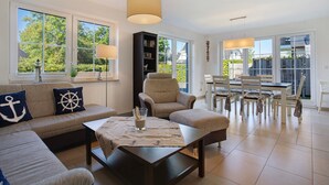 A living room with a sofa, armchair, dining table, and chairs, large windows, and a bookshelf. - Holiday Home 'Ferienhaus Strandperle' with Private Terrace, Private Garden and Wi-Fi (Ostseebad Zingst)