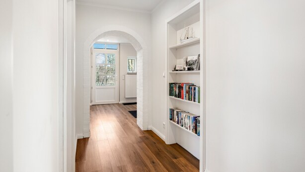 Un corridoio con pavimento in legno, pareti bianche e una libreria con libri e oggetti decorativi.