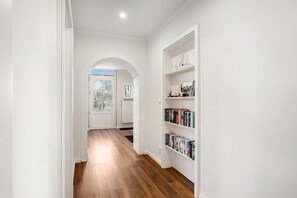 Un pasillo con suelo de madera, paredes blancas y una estantería con libros y objetos decorativos.