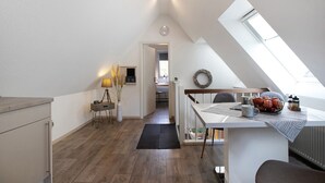 A loft-style room with a sloped ceiling, a dining area with a table and chairs, a small kitchen area, and a view of the outdoors. - Apartment 'Kleine Nock' with Balcony and Wi-Fi (Burg (Dithmarschen))