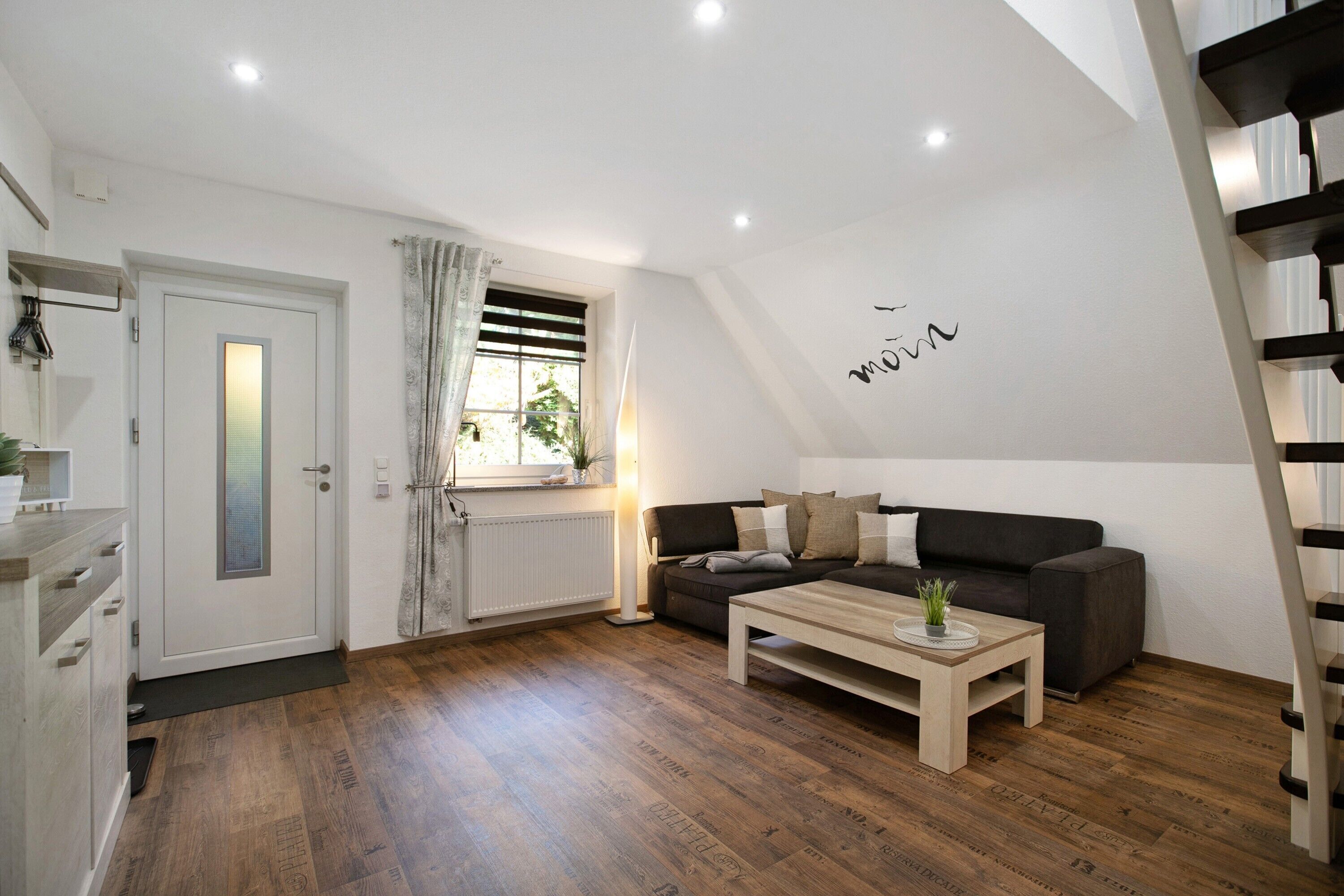 A modern living room with a wooden floor, a black sofa, a wooden coffee table, and a staircase.