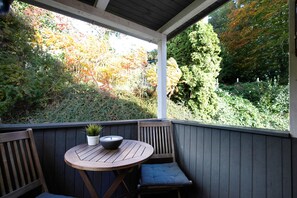 Eine Holzterrasse mit einem runden Tisch und Stühlen, einem Blumentopf und Blick auf Bäume und Grünpflanzen.