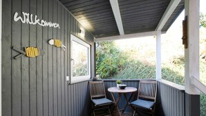A wooden deck with two chairs and a small table, a window, and a 'Willkommen' sign on the wall. - Apartment 'Kleine Nock' with Balcony and Wi-Fi (Burg (Dithmarschen))