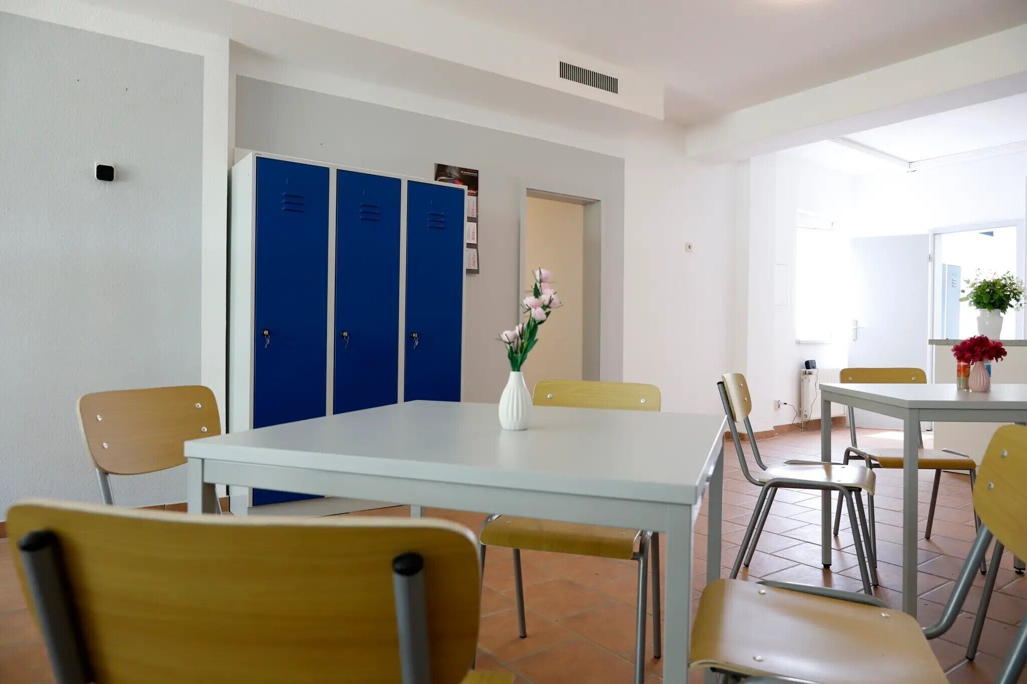 A room with a white table, yellow chairs, blue lockers, and a vase with flowers.