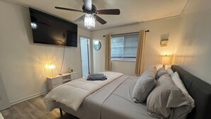A bedroom with a bed, a ceiling fan, a TV mounted on the wall, a bedside table with a lamp, and a window with curtains. - Cozy 4BR Home with Office, Fitness Area, Spacious Backyard & Pet-Friendl (Monroe)