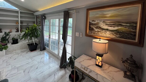 A spacious room with a marble floor, a large window with curtains, a painting of a seascape, and a table with a lamp and decorative items. - Cozy 4BR Home with Office, Fitness Area, Spacious Backyard & Pet-Friendl (Monroe)