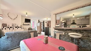 Una sala de estar con sofá, una mesa de comedor con un jarrón y una zona de cocina con pared de piedra y televisor.