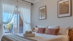 A neatly made bed with a striped blanket, two pillows, and rolled towels. There are two framed artworks on the wall, a window with sheer curtains, and a small bedside table with a lamp. - Sunset, Apartment in Costa Adeje - Relax in Paradise (Costa Adeje)