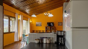 Una cocina con encimera negra, refrigerador blanco y cafetera. Hay frascos sobre la encimera, un televisor en la pared y una ventana con cortinas.