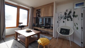 A modern kitchen with wooden cabinets, a dining table, a TV, and a white chair with a leaf-patterned wall. - Apartment 'La Grange' with Mountain View, Private Garden and Wi-Fi (Cazeaux-de-Larboust)