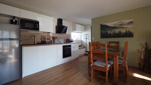A modern kitchen with white cabinets, a microwave, and an oven. There is a dining area with wooden chairs and a table. - Apartment 'La Grange' with Mountain View, Private Garden and Wi-Fi (Cazeaux-de-Larboust)