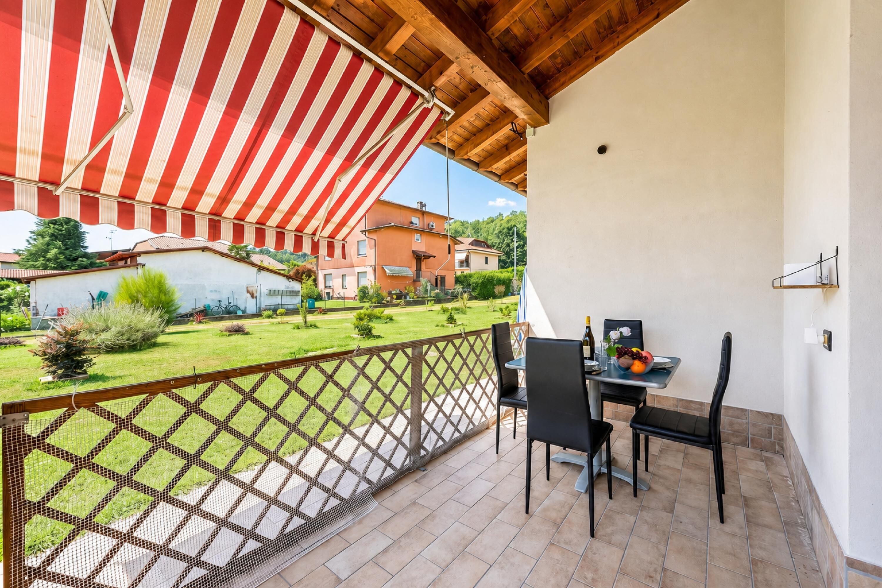 Un balcon doté d’un auvent rayé de rouge et de blanc, une table avec des chaises, et une vue sur un jardin et des bâtiments.