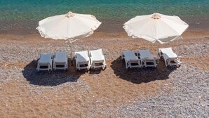 Una playa con sillas de descanso blancas y sombrillas, un velero y agua azul cristalina.