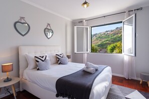 Ein Schlafzimmer mit einem großen Bett, zwei herzförmigen Spiegeln, Blick auf eine Hügellandschaft und einem Fenster mit Vorhängen.