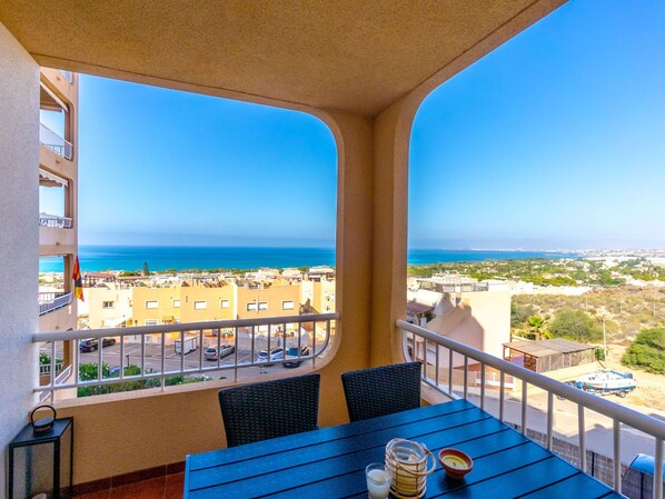 A balcony with a table and chairs, overlooking a coastal town and the sea.