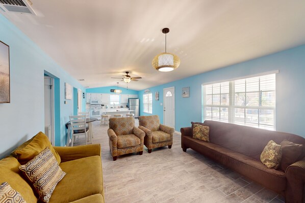 A spacious living room with a brown sofa, two patterned armchairs, and a yellow couch. A kitchen is visible in the background with a dining area.