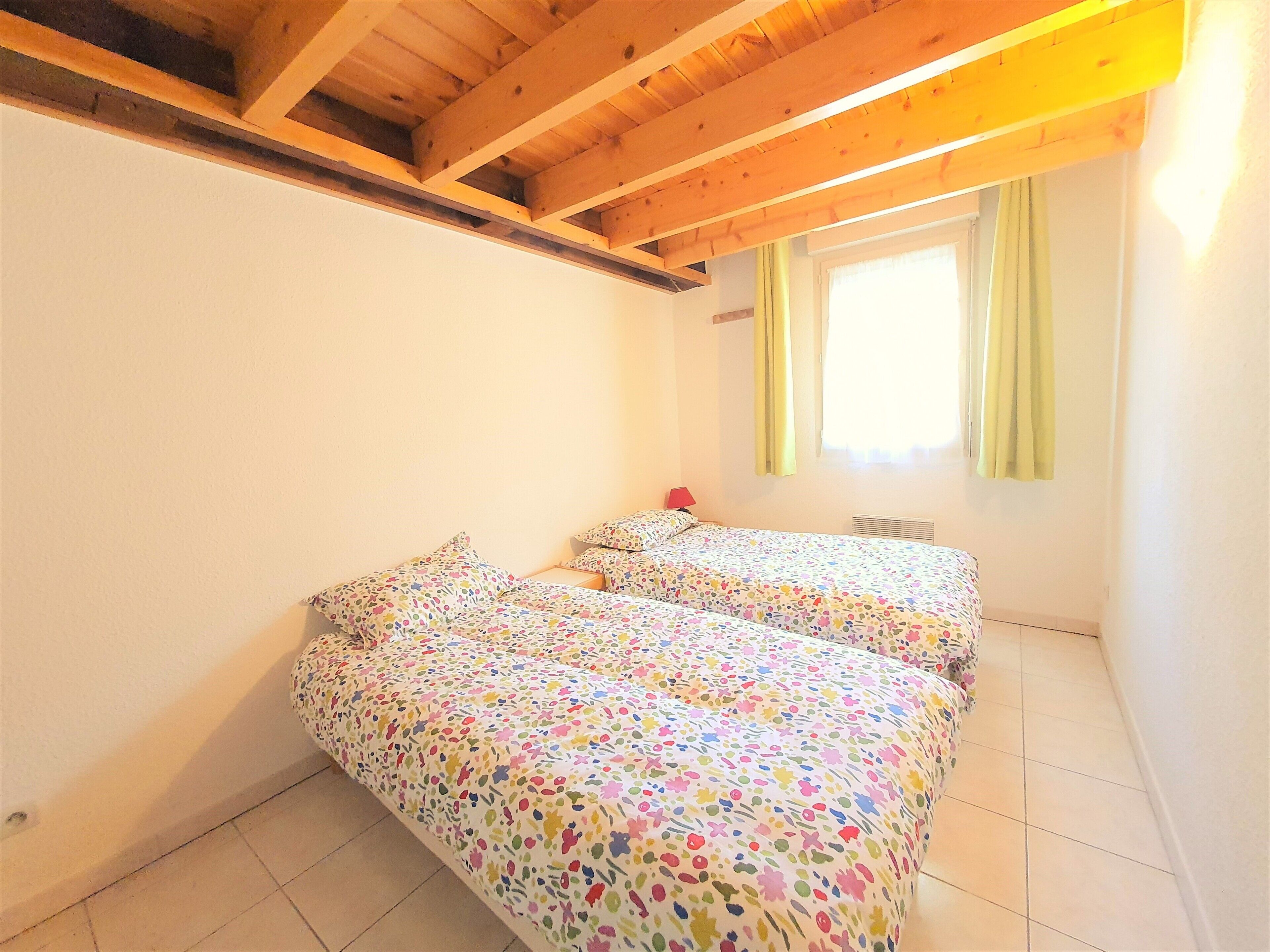 A room with two beds, wooden ceiling beams, a window with curtains, and a radiator.