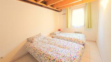 A room with two beds, wooden ceiling beams, a window with curtains, and a radiator.