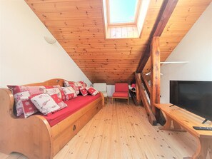 Ein gemütliches Dachzimmer mit Holzbalkendecke, einem roten Sofa mit gemusterten Kissen, einem kleinen roten Sessel, einem Fernseher auf einem Holzschrank und einem Oberlicht.