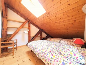 A room with a sloped ceiling, wooden beams, a bed with patterned bedding, a chair, and a small table.