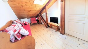 Kodikas ullakkohuone, jossa on kalteva katto, puiset kattopalkit, pienelle pöydälle sijoitettu televisio, punainen tuoli ja kuviollinen vuodevaatteilla varustettu sänky.
