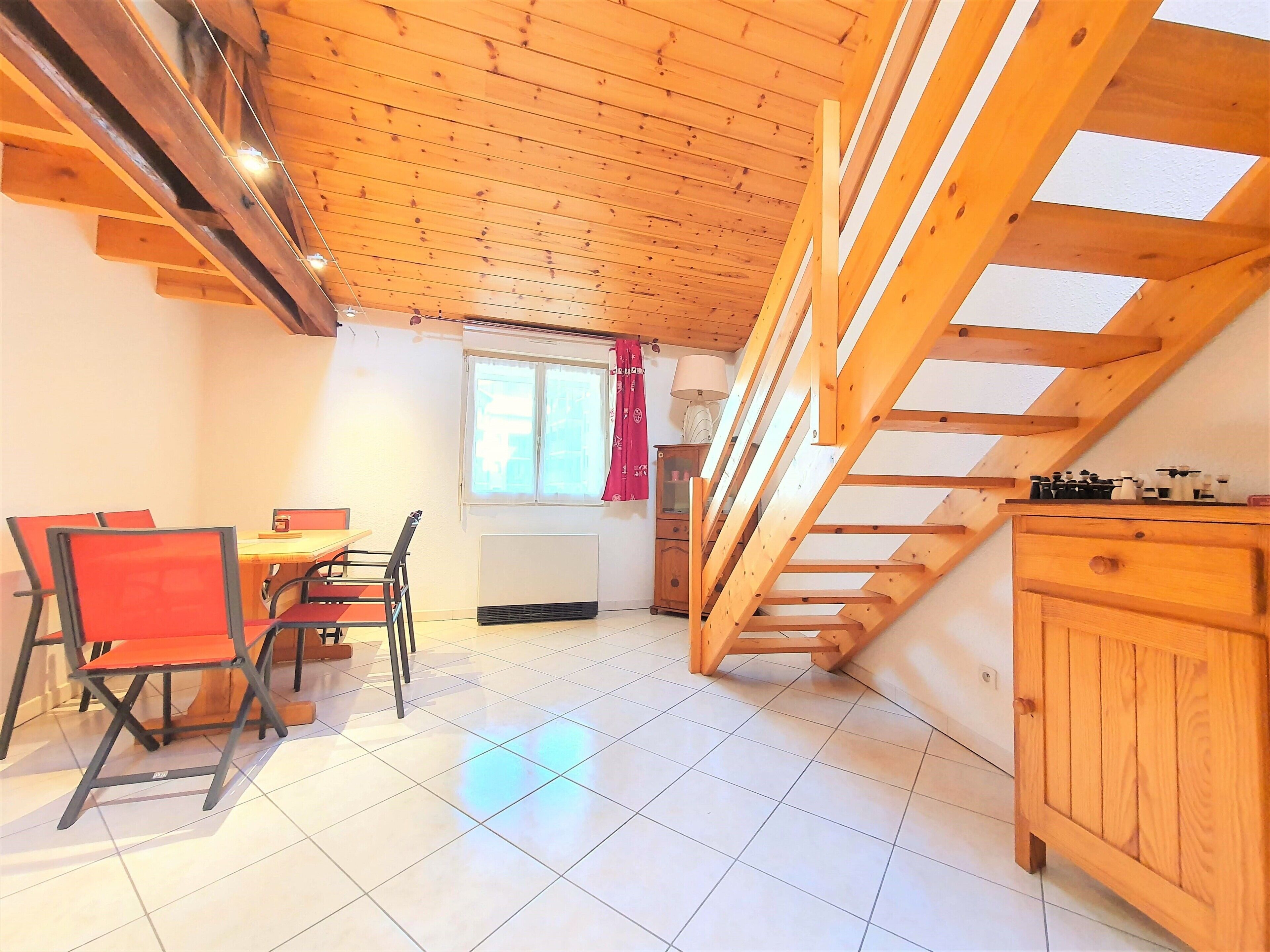 Een kamer met een schuine kap, een eettafel met stoelen, een raam met gordijnen en een trap.