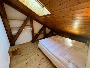 A small attic bedroom with a sloped ceiling, wooden beams, a single bed, a small table, and a skylight.