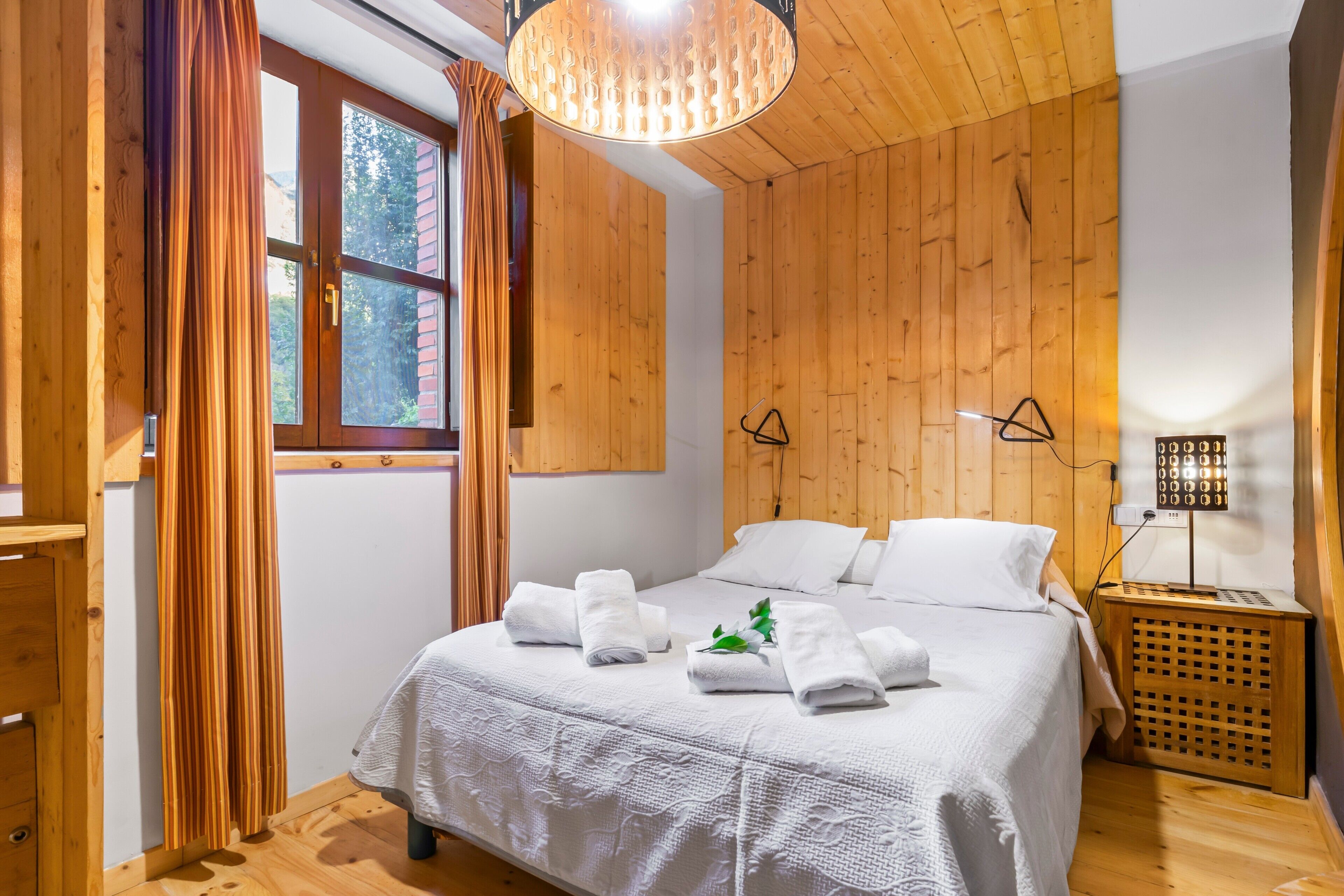 Un dormitorio con paneles de madera, una cama, sábanas blancas, una lámpara colgante y una mesita de noche con lámpara.