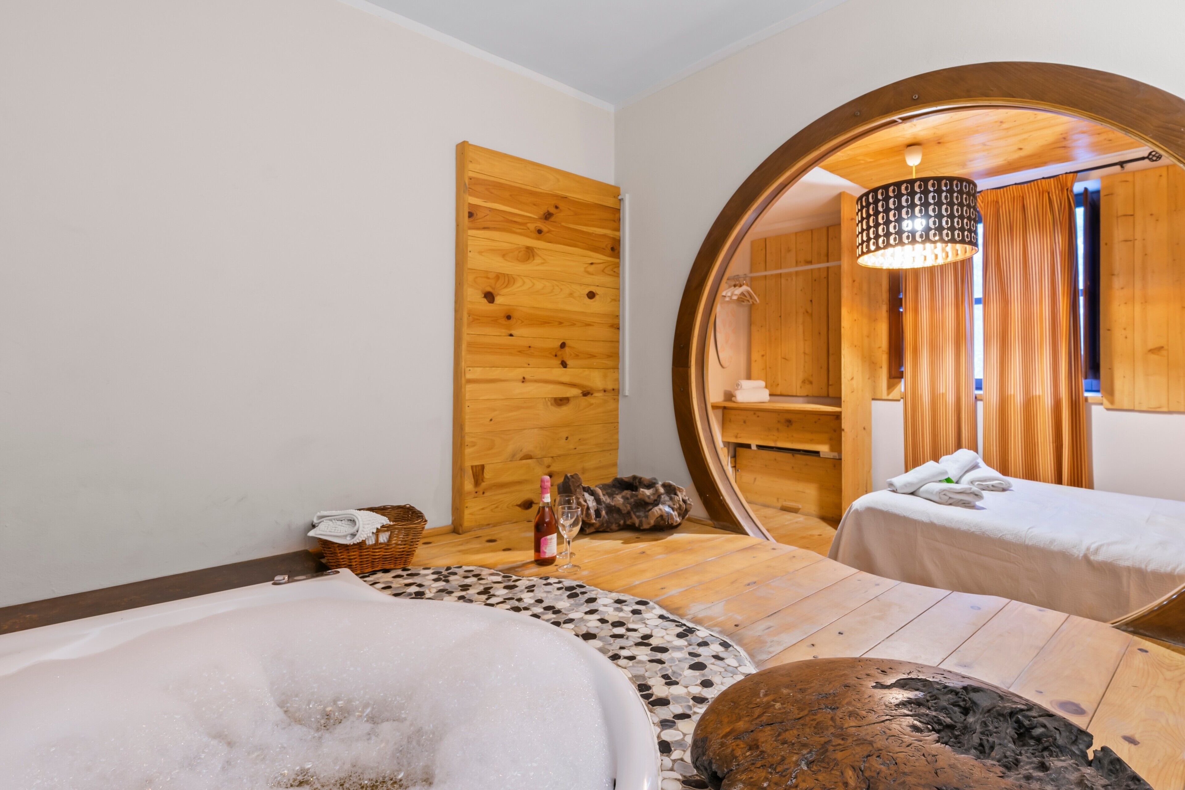 A spa room with a wooden floor, a round mirror, a bathtub, a massage table, and a bottle of wine.