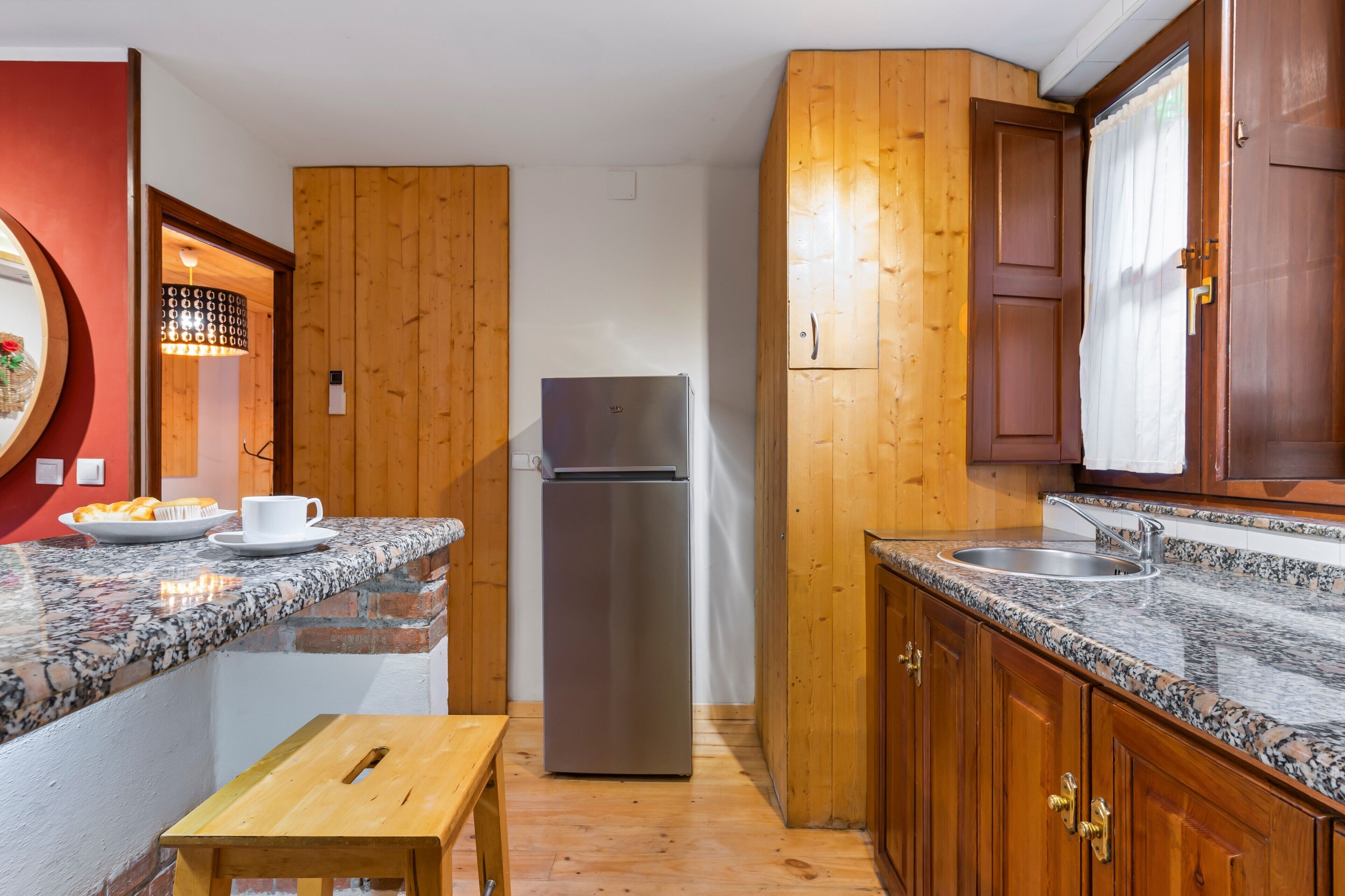 A compact kitchen with wooden cabinets, a stainless steel refrigerator, granite countertops, a wooden stool, and a small window with curtains.