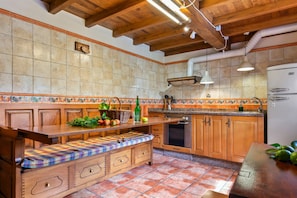 Uma cozinha com armários de madeira, um banco com almofada xadrez, uma geladeira e um painel de azulejos.
