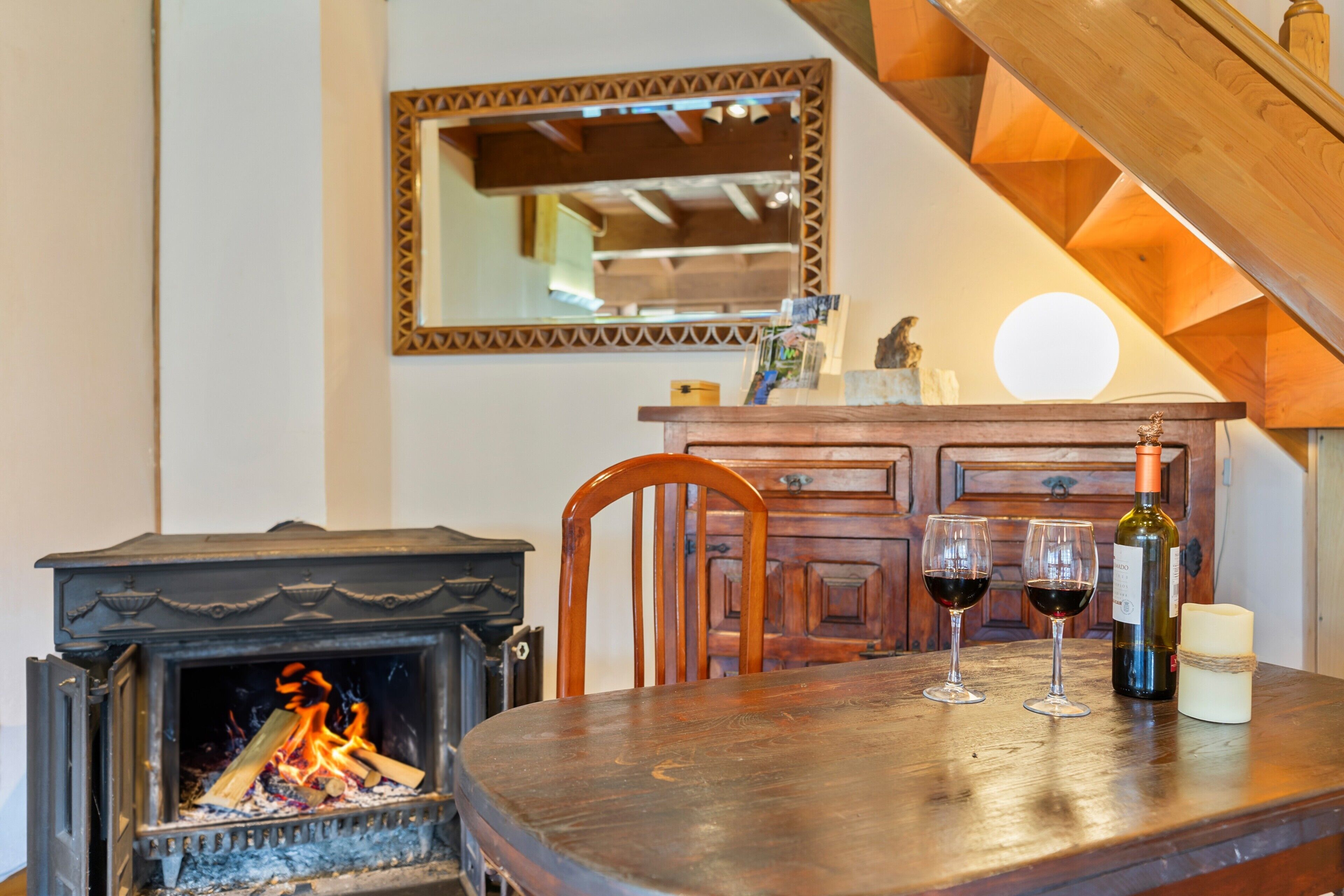 Una mesa de madera para comer con dos copas de vino y una vela, una chimenea, una silla de madera y un espejo en la pared.