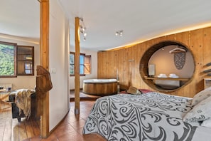 A room with a wooden interior, a round mirror, a bed with patterned bedding, a wooden bathtub, and a window with a view of greenery. - Hot Tub with Mountain Views in Ponga Natural Park (Cazo)