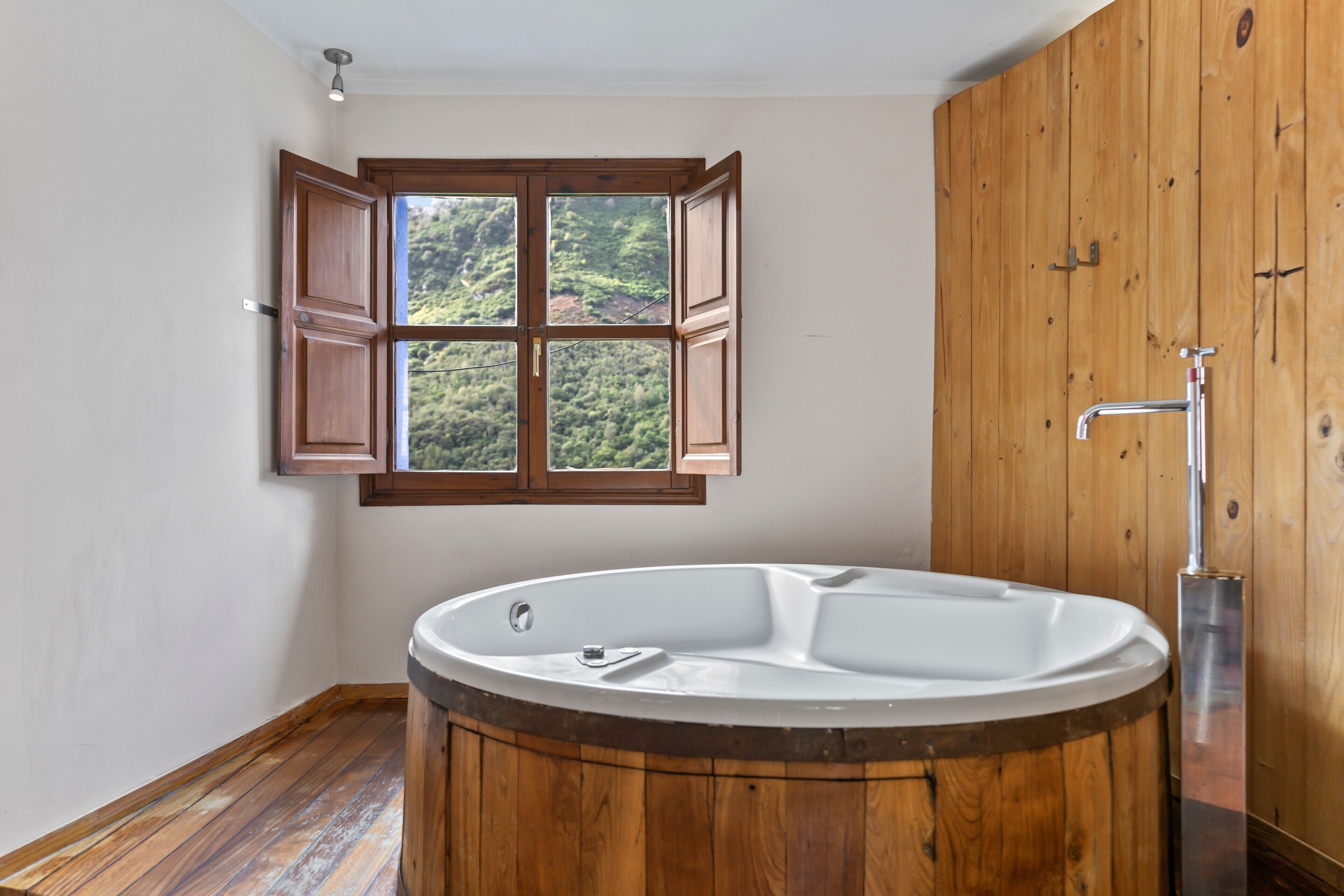 Een badkamer met een houten bad, een raam met open luiken en een houten deur.