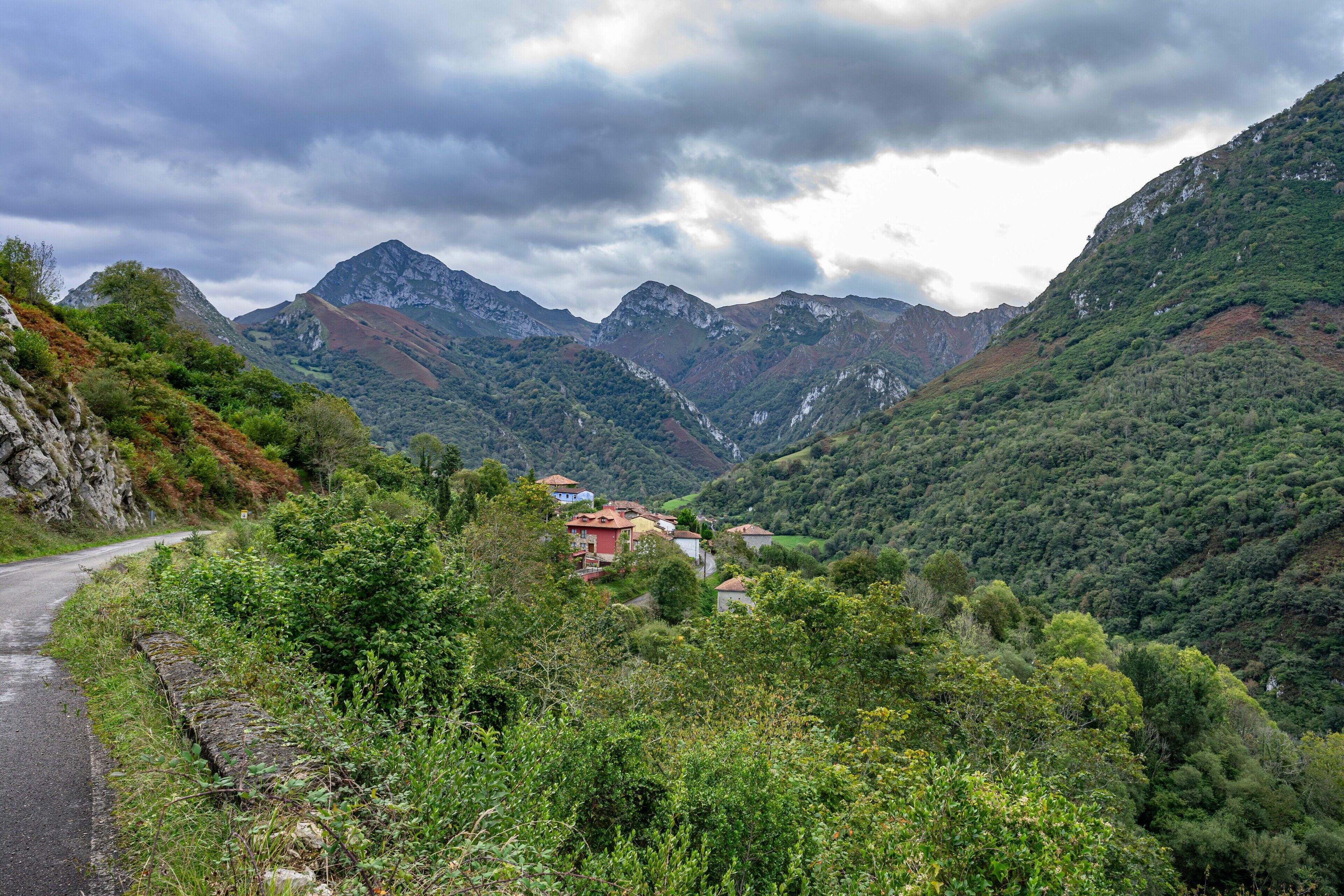 Paisagem montanhosa com estrada sinuosa, pequena aldeia com casas tradicionais e um muro de pedra à esquerda.