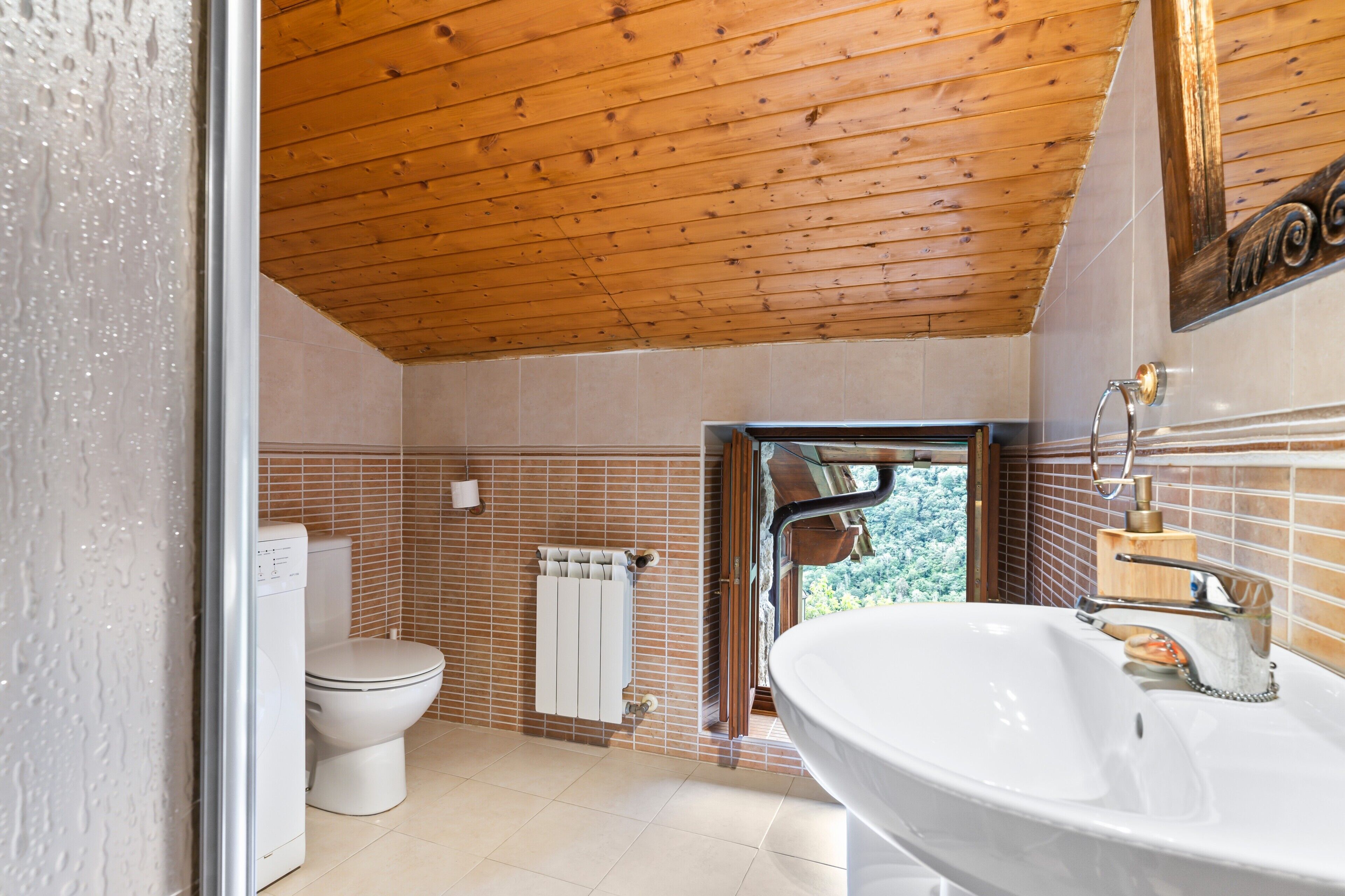 Un baño con techo de madera, inodoro y lavamanos blancos, y vistas a la vegetación exterior.