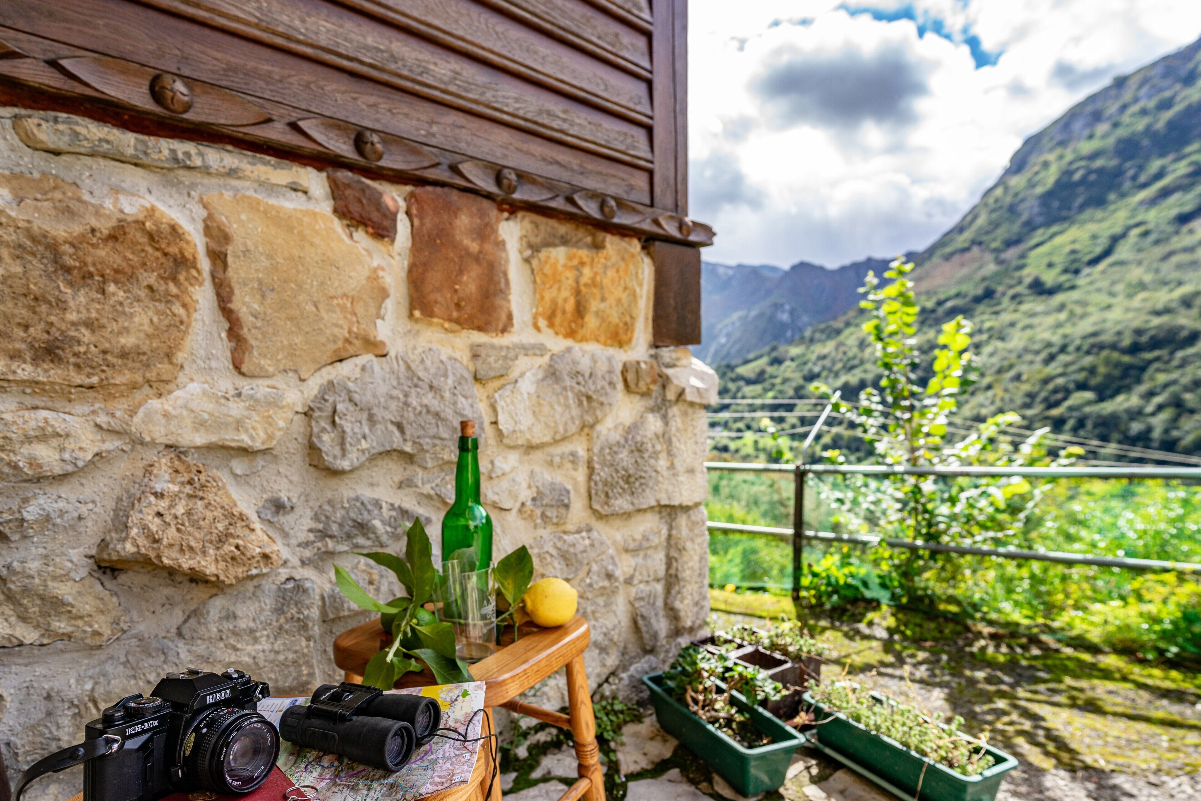 A stone wall, a wooden table with a camera, a bottle, a glass, and a lemon.