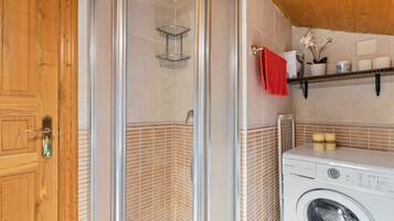 Un rincón de una habitación con una puerta de madera, un box de ducha de vidrio, una lavadora y un estante con artículos de aseo.