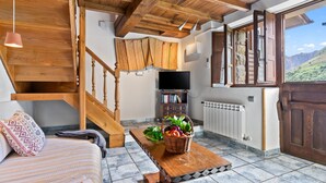 A rustic interior with a wooden staircase, a flat-screen TV, a bookshelf, and a window with a mountain view. - El Rincón del Sol: Retreat and Rest in the Picos de Europa. (Cazo)