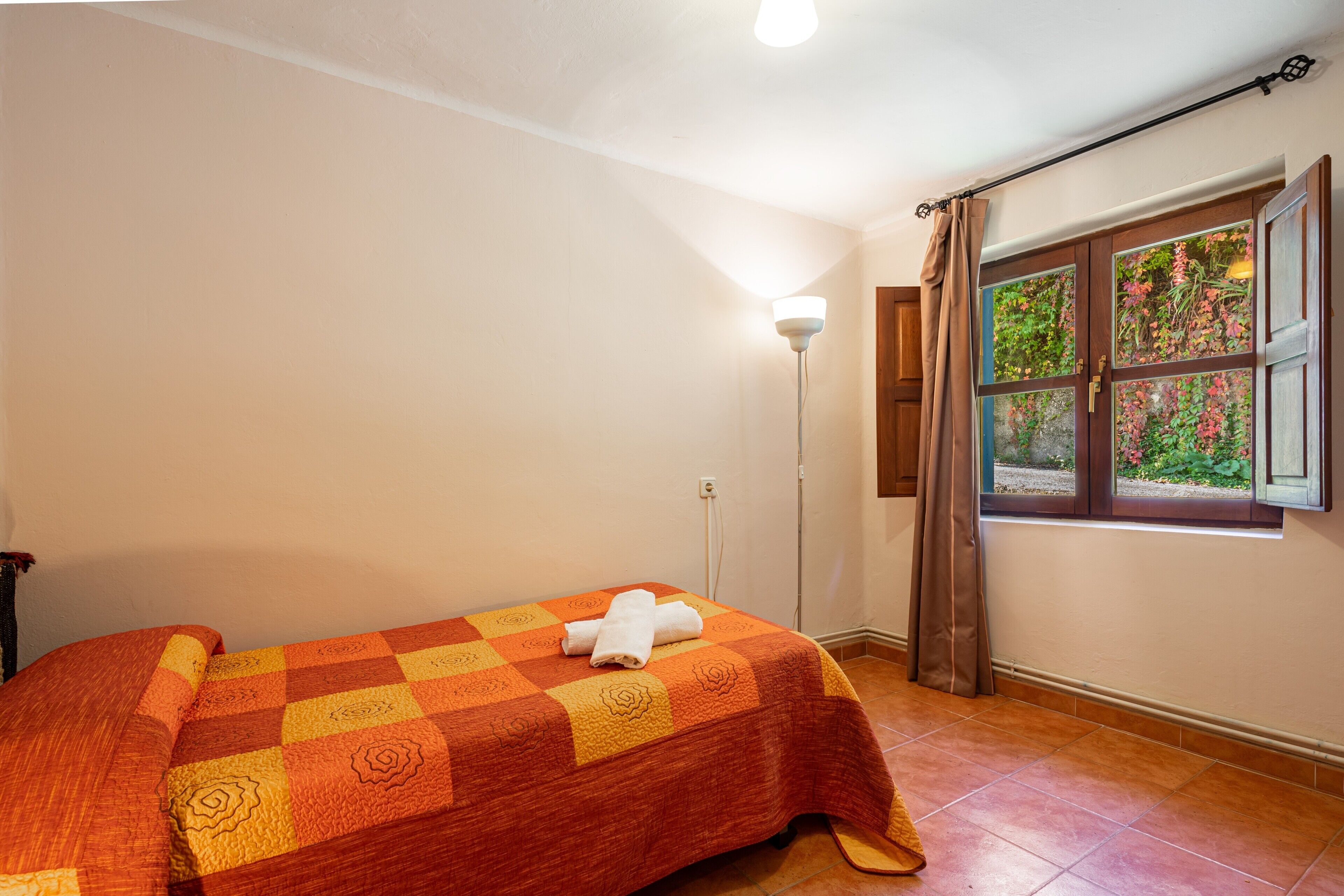 A room with a bed, a window with curtains, a floor lamp, and a view of greenery outside.