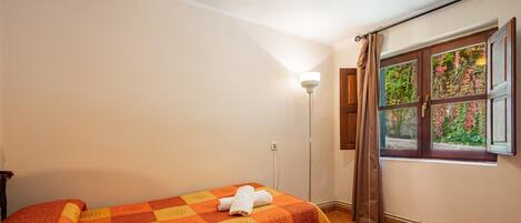 A room with a bed, a window with curtains, a floor lamp, and a view of greenery outside.