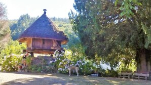 A rustic thatched-roof structure with a wooden staircase leading up to it, surrounded by lush greenery and flowering plants. - Rural House 'Teixo' with Mountain Views, Private Terrace, and Wi-Fi (A Fonsagrada)