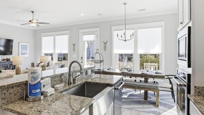 A modern kitchen with granite countertops, stainless steel appliances, and a large island. There is a dining area with a table and chairs, and a view of the outside through large windows. - Park's Edge Home | Pool, Bike Trails, Beach Access | Three Seas Villa by Kaiser (Orange Beach)