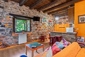 A rustic interior with stone walls, wooden beams, a stone fireplace, a wooden table, and a sofa with colorful cushions.