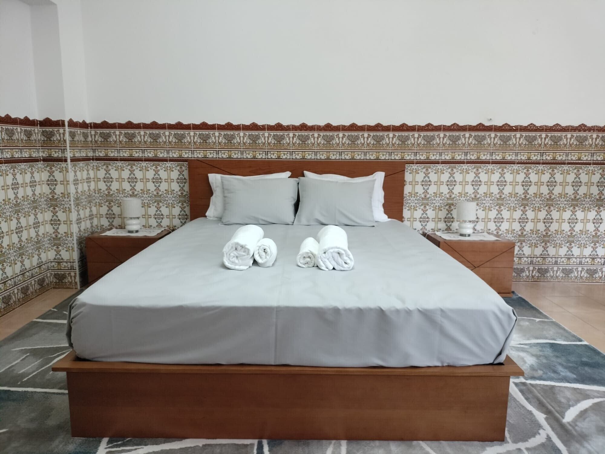 Een net opgemaakt bed met kussens, een nachtkastje met een lamp, en een behangen muur.