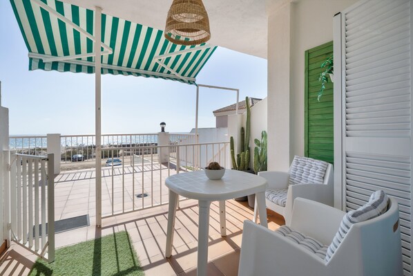 Un balcon avec une table blanche, des chaises, un auvent à rayures et une vue sur la mer.