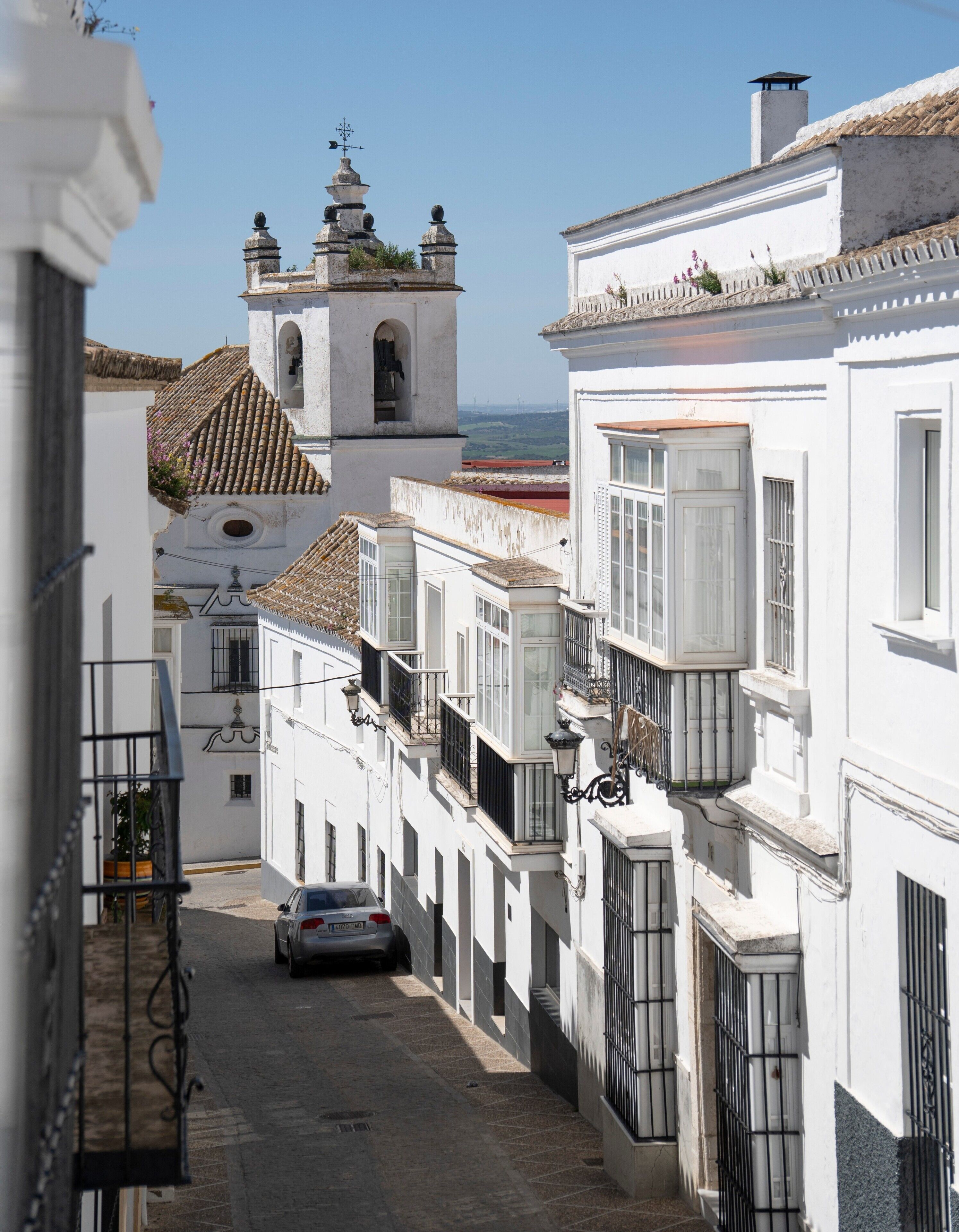 A narrow street with white buildings, a bell tower, and a car parked on the street.