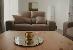 Uma mesa de madeira redonda com um tabuleiro dourado e uma tigela espelhada. Ao fundo, um sofá castanho e uma porta branca.